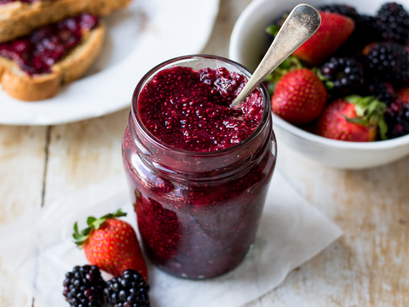 Image of dark berry chia jam in small curved jar, copper spoon in the jar, berries in background