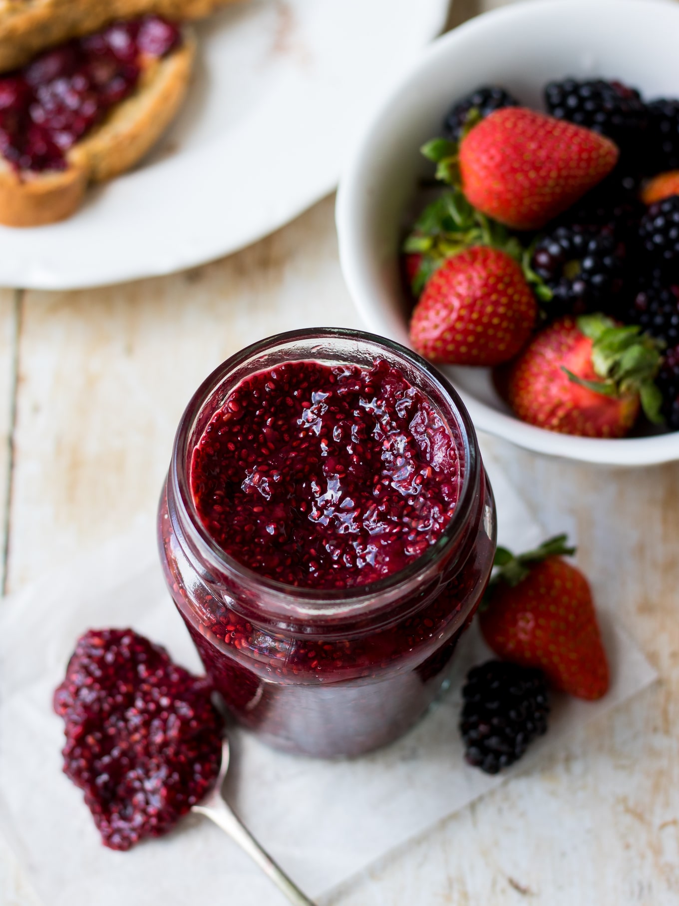 Image of berry chia jam in a small glass jar, shot from a side angle, with a bowl of mixed berries in background.