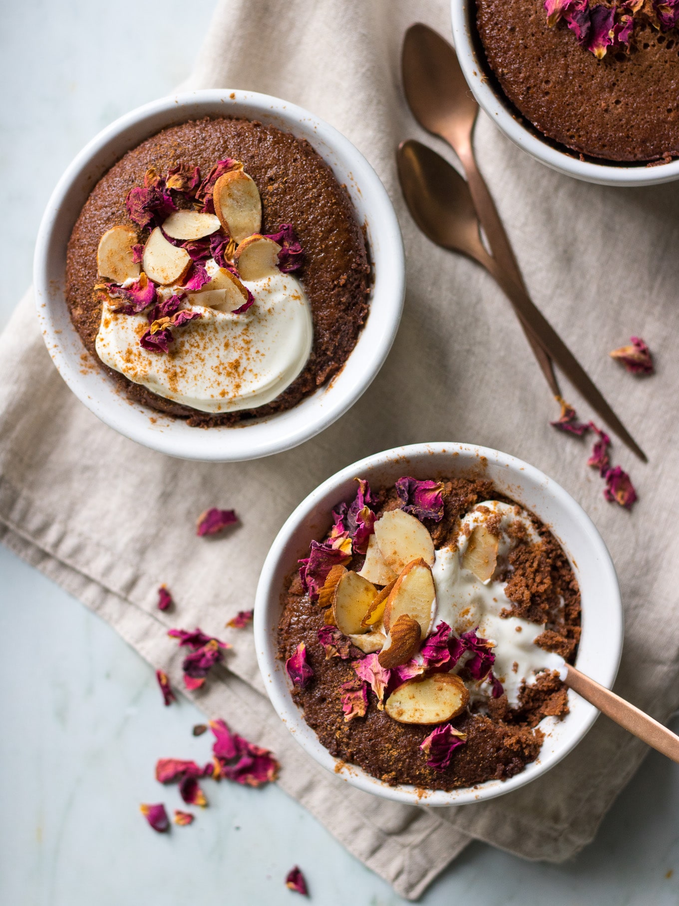 A healthy chocolate cake served in a white ramekin with rose petals, almonds and yoghurt