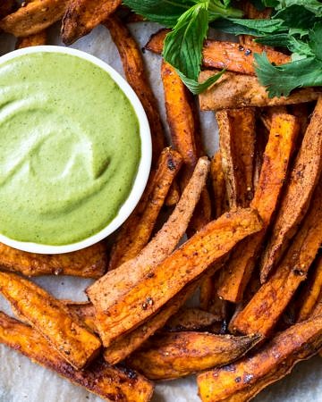 Cinnamon Paprika Sweet Potato Fries arranged on a plate with dipping sauce and herbs.