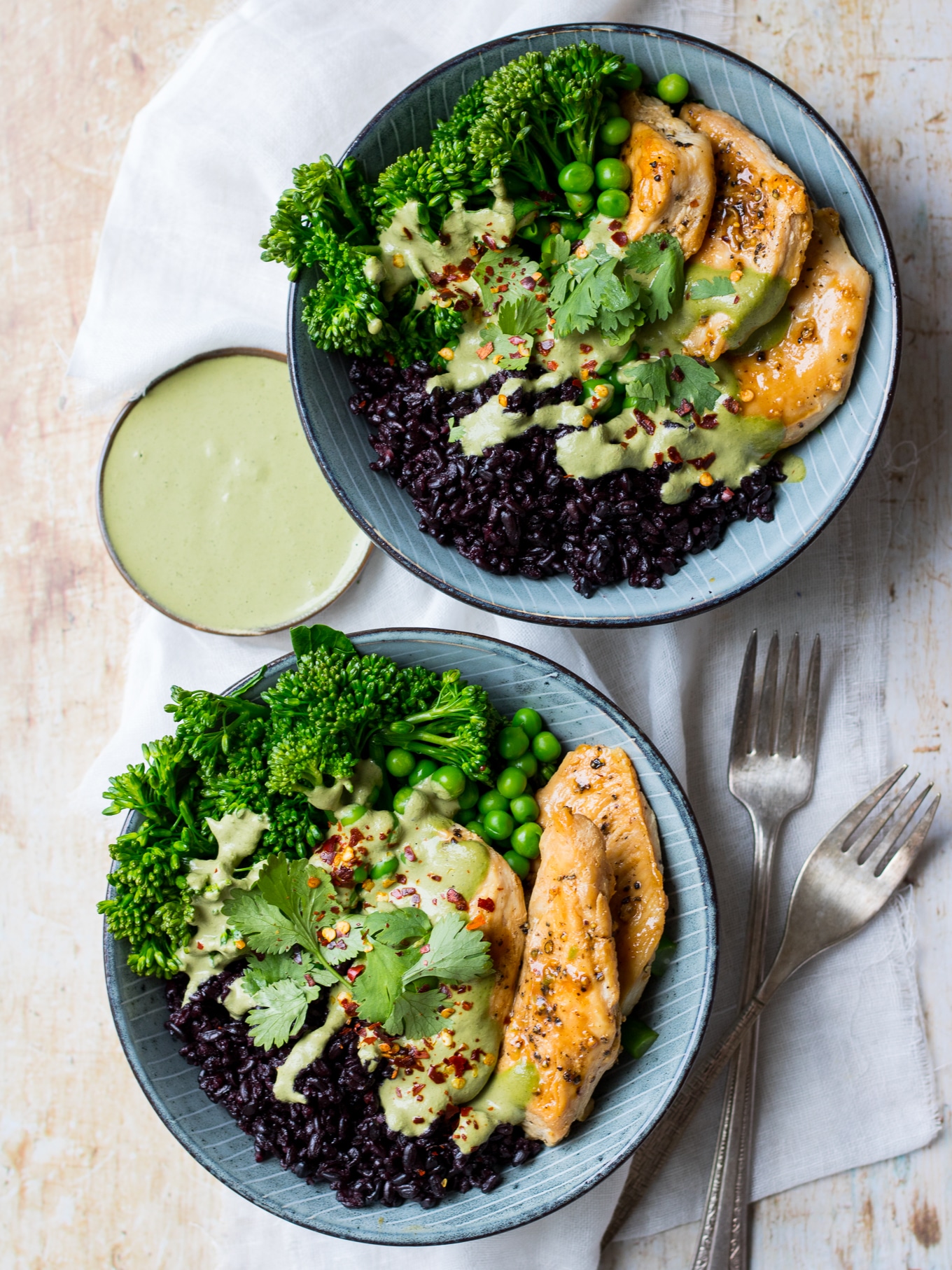 Chicken black rice bowls; two blue bowls filled with black rice, chicken and broccolini, side of green herb sauce, on a light table