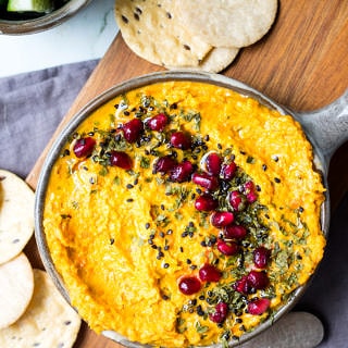 Roasted carrot tahini dip in grey serving bowl topped with sesame seeds and pomegranate and herbs, on chopping board.