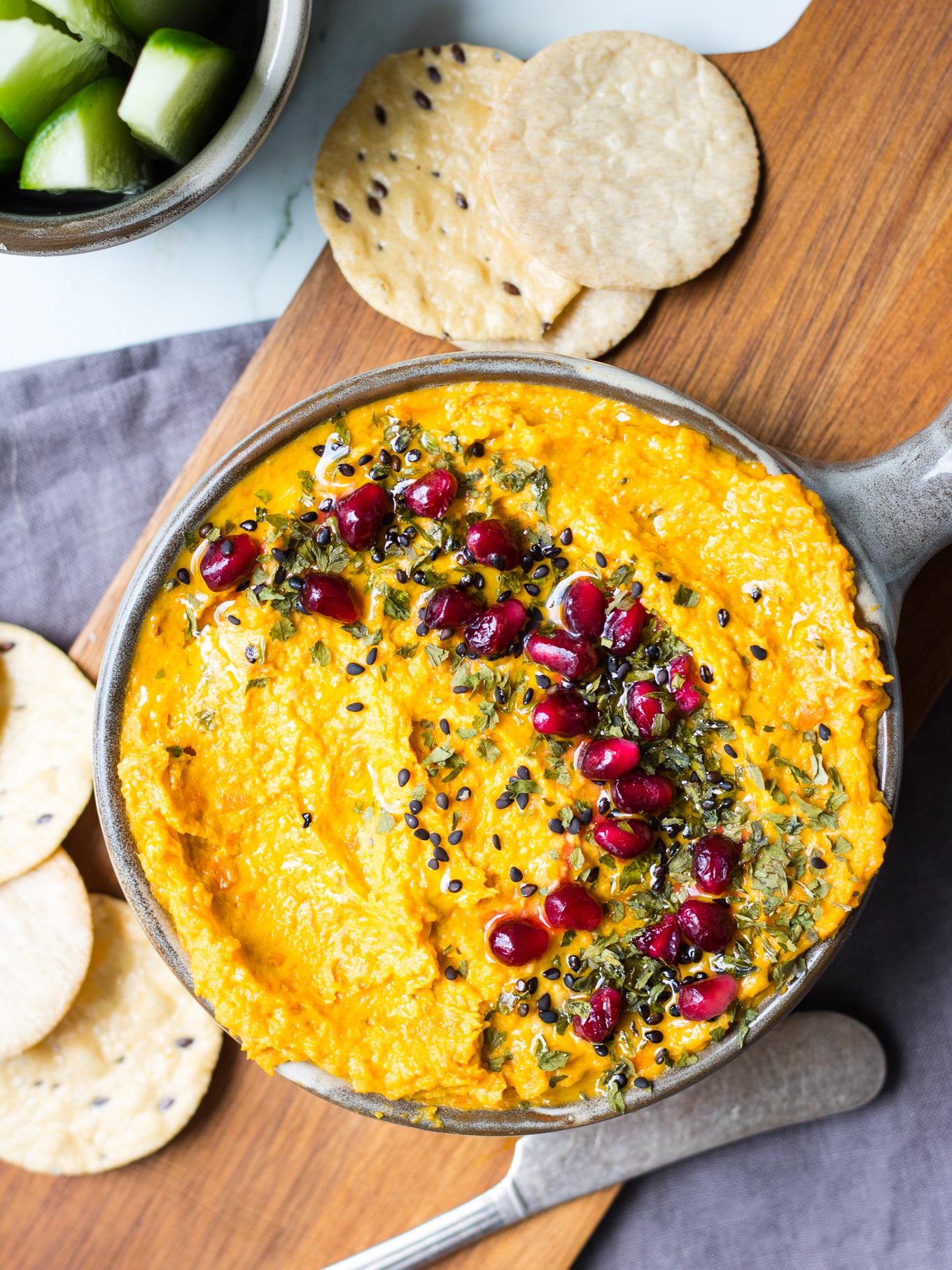 Roasted carrot tahini dip in grey serving bowl topped with sesame seeds and pomegranate and herbs, on chopping board.