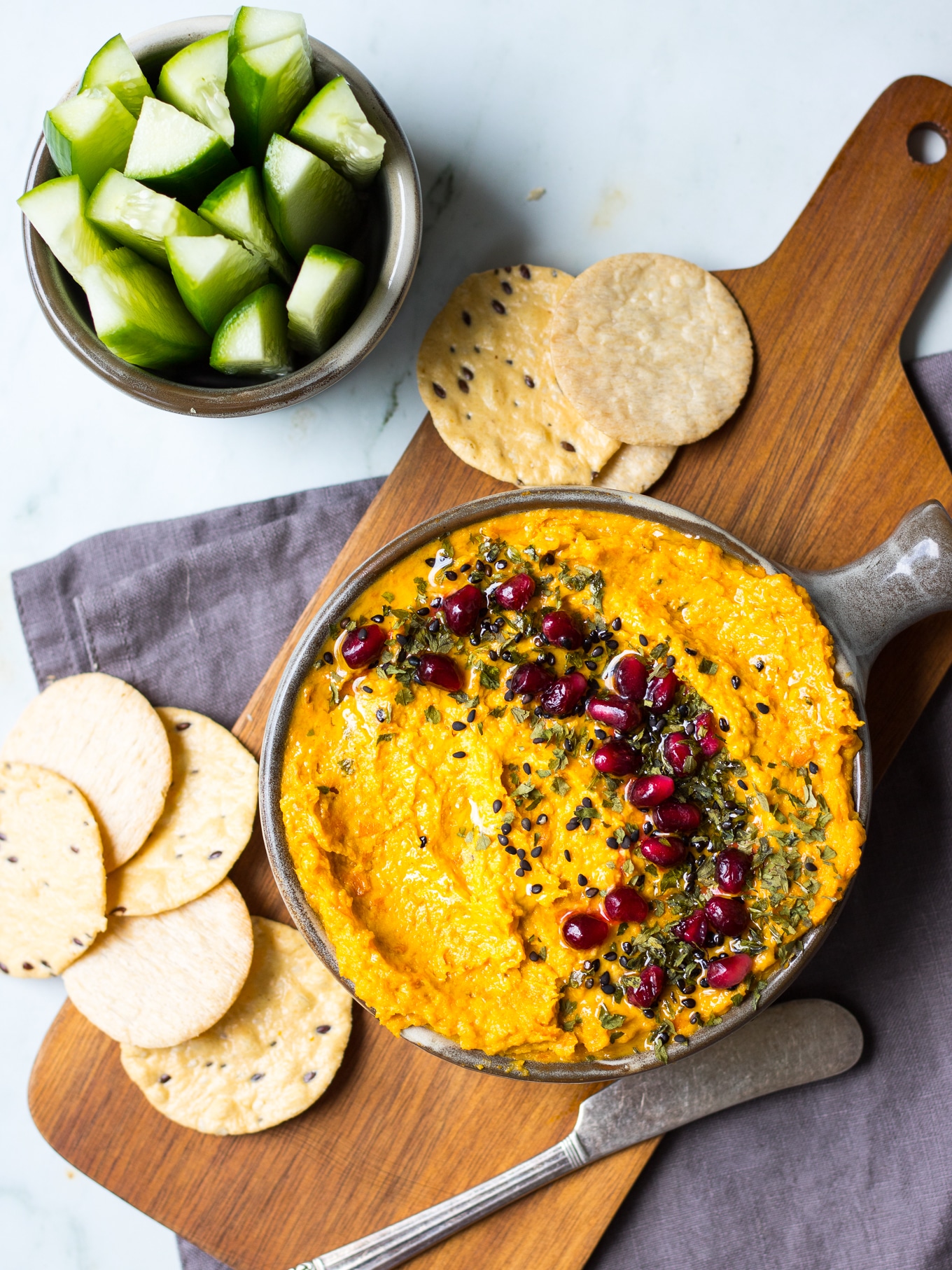 Roasted carrot tahini dip in grey serving bowl topped with sesame, pomegranate and herbs, on chopping board. With crackers.