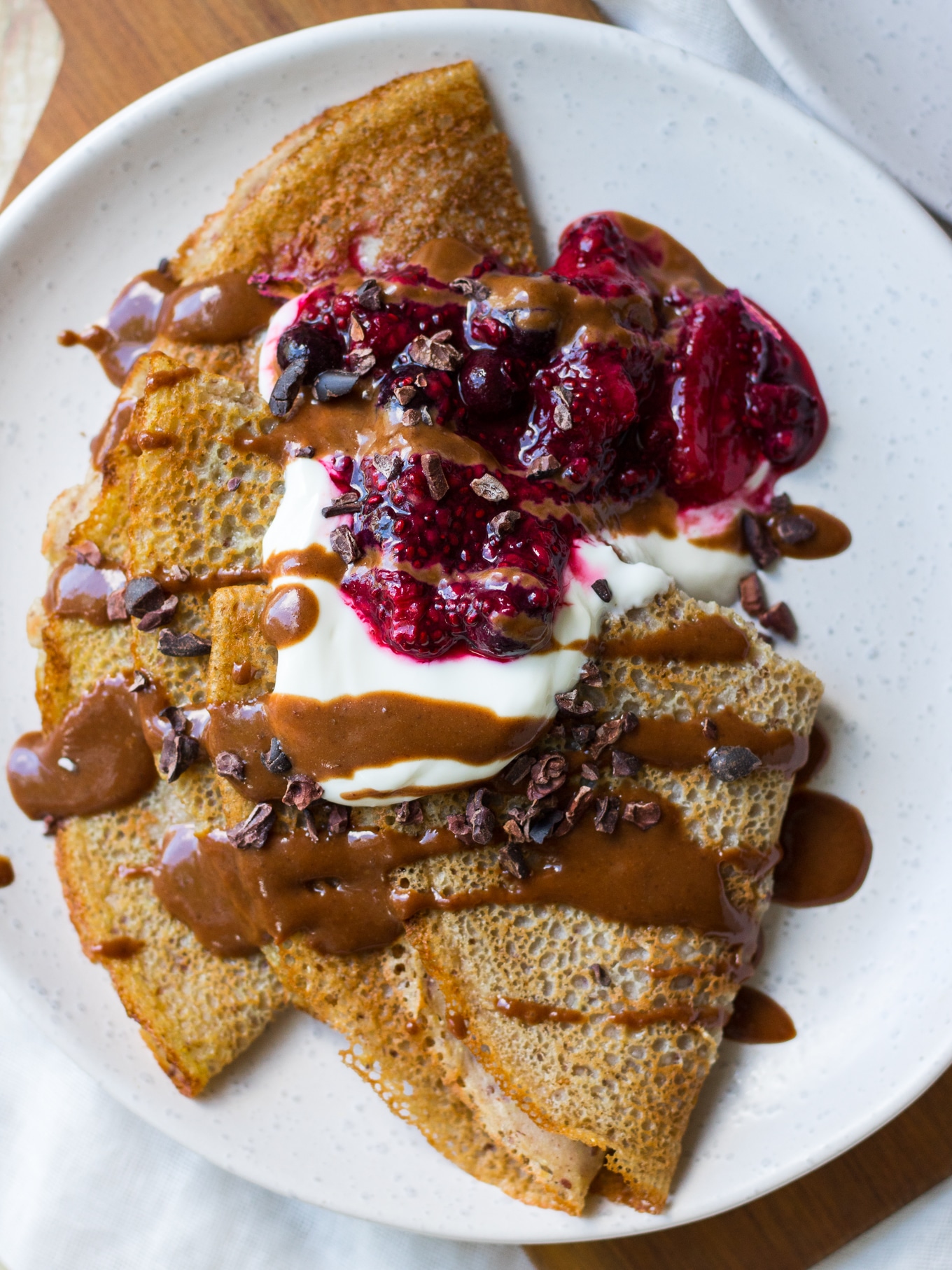 Sweet Vegan Buckwheat Crepes, folded on a white plate, topped with yoghurt, berry sauce, chocolate drizzle.