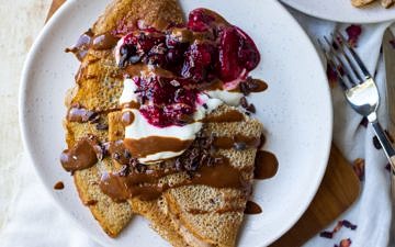 Sweet vegan buckwheat crepes folded on white plate served with yoghurt, berries and chocolate sauce.