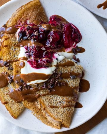 Sweet vegan buckwheat crepes folded on white plate served with yoghurt, berries and chocolate sauce.