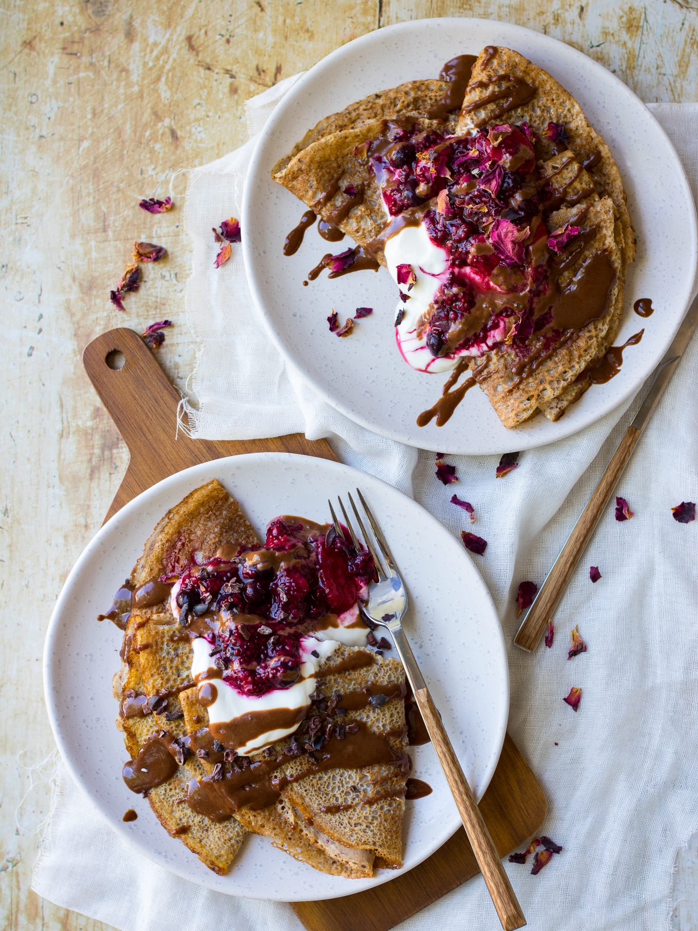 Two plates of vegan buckwheat crepes. Folded crepes with yoghurt, berry sauce and chocolate sauce drizzle.