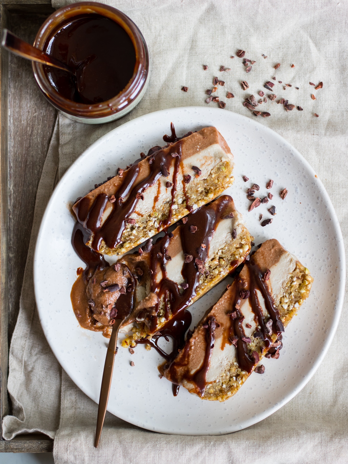 Vegan chocolate banana nice cream cake, three slices on a cream plate, chocolate sauce drizzled on top, cacao nibs scattered
