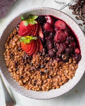 Chocolate buckwheat porridge in patterned bowl on marble background, spoon, berries, cacao nibs