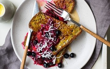 Golden milk french toast stacked on a white plate topped with berry compote and flaked coconut.