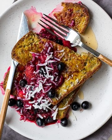 Golden milk french toast stacked on a white plate topped with berry compote and flaked coconut.