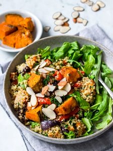 Honey roast pumpkin quinoa salad in grey bowl with rocket, flaked almonds on top, roast pumpkin in background