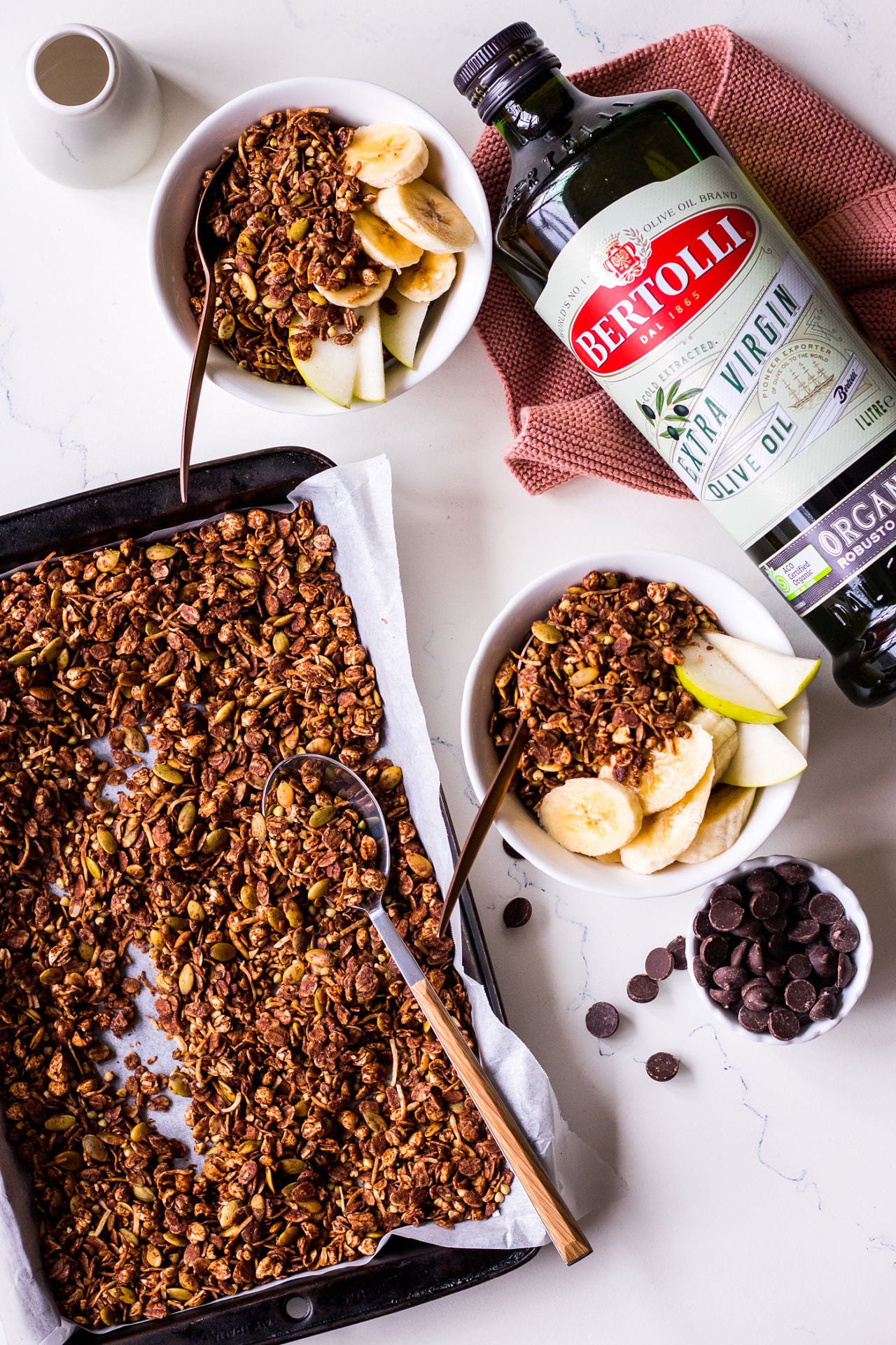 Bottle of Bertolli Organic Extra Virgin Olive Oil laid next to a baking tray and two small bowls with fruit and chocolate granola