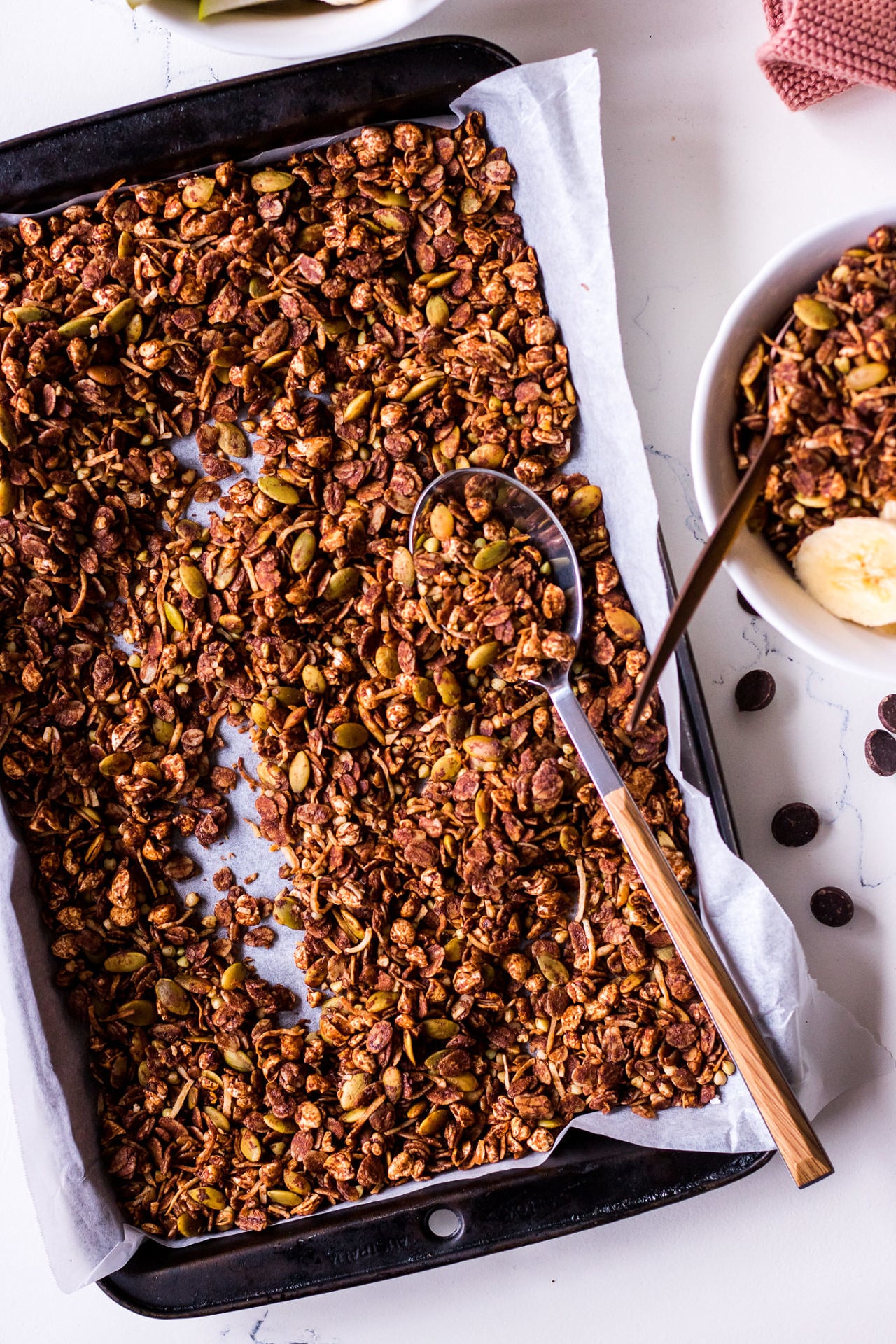 Baking tray filled with chocolate olive oil granola with a wooden handled spoon, small white cereal bowl to the side