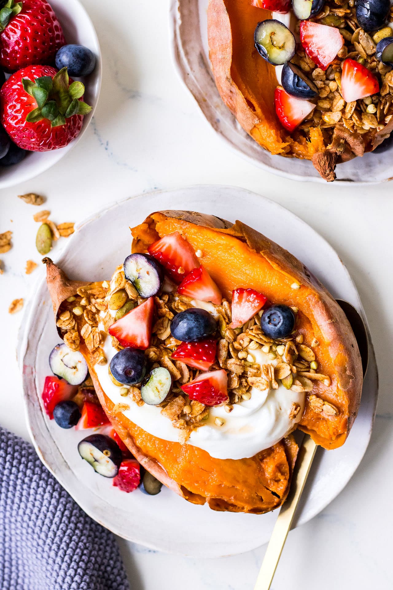 Small white plate with open sweet potato topped with yoghurt, strawberries, blueberries and oat granola, gold spoon to side