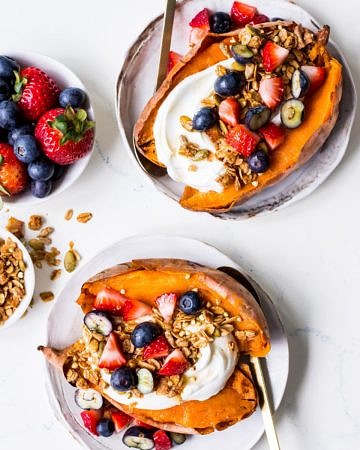 Top view of two white plates of breakfast sweet potatoes topped with yoghurt, berries and granola, with gold spoons