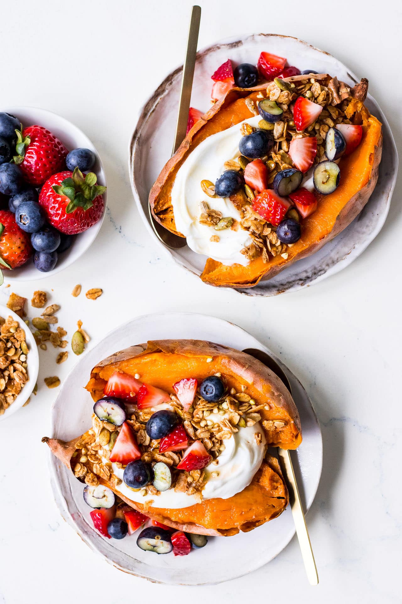 Top view of two white plates of breakfast sweet potatoes topped with yoghurt, berries and granola, with gold spoons