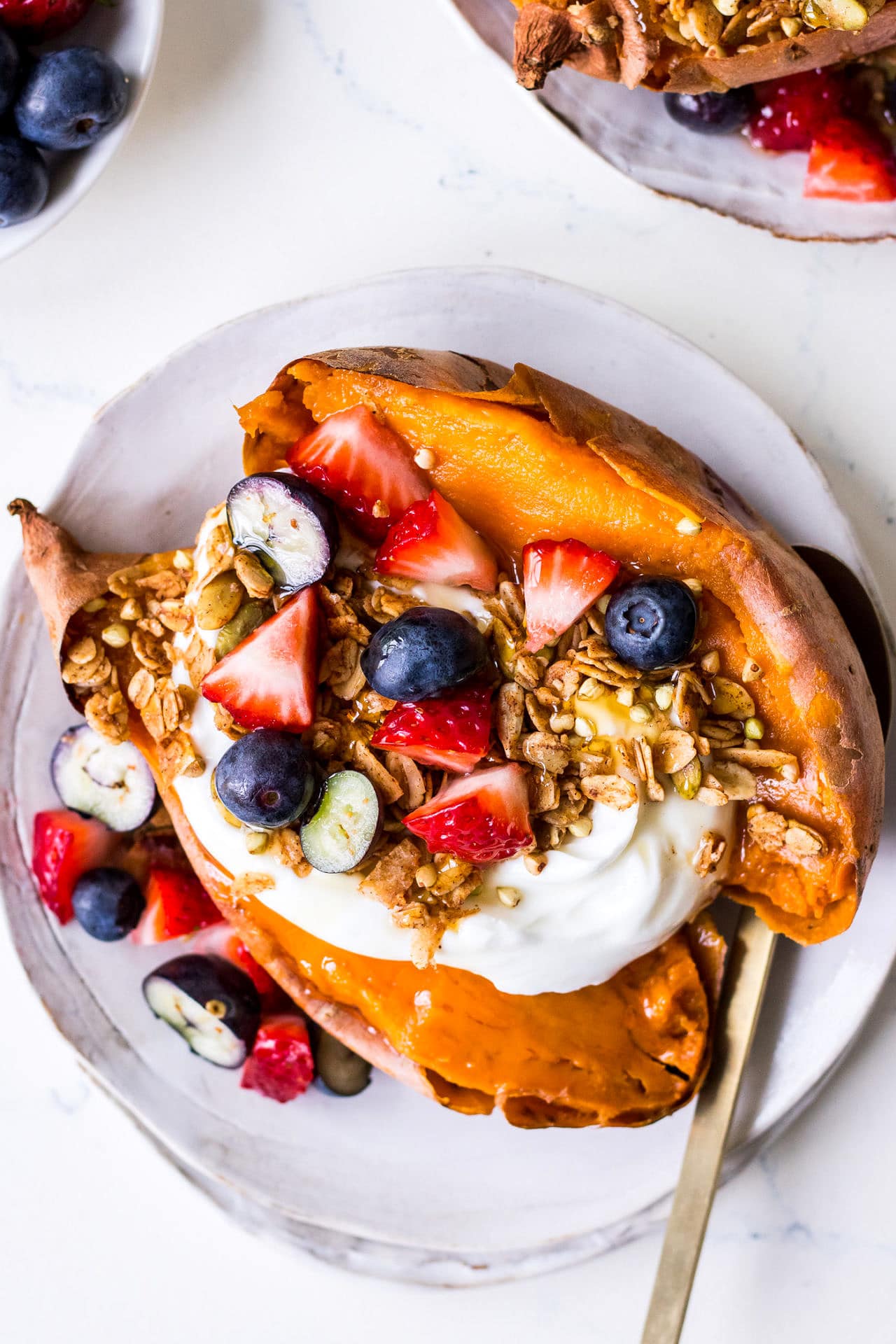 Close up of sweet potato halved and topped with Greek yoghurt, sliced berries, oat granola, gold teaspoon to side, on a white side plate