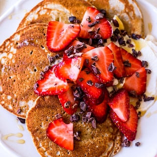 Close up of buckwheat pancakes layered on a white plate with sliced strawberries and cacao nibs sprinkled on top