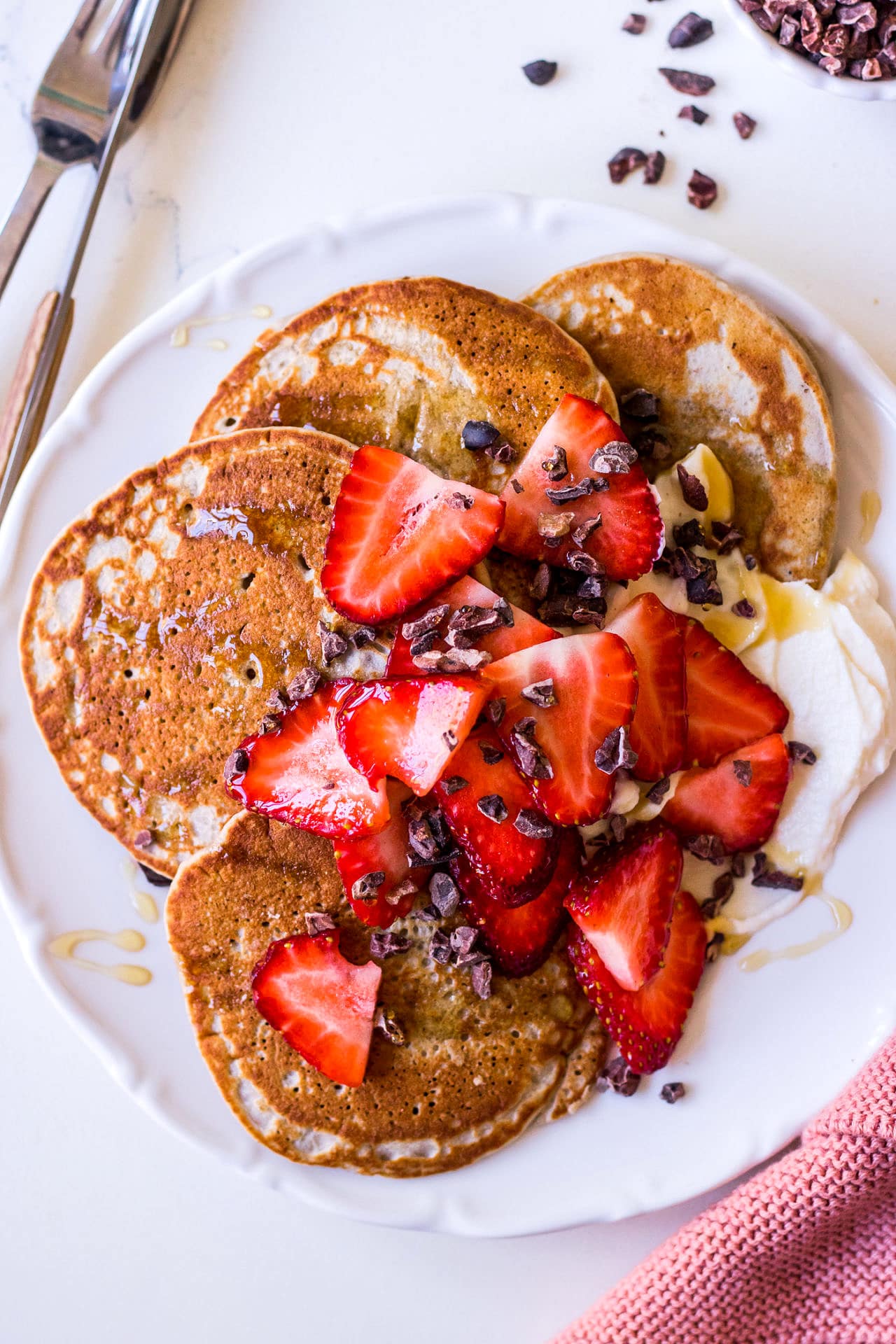 Piatto rotondo bianco, pancake proteici di grano saraceno a strati marrone dorato, fragole a fette, pennini di cacao sparsi