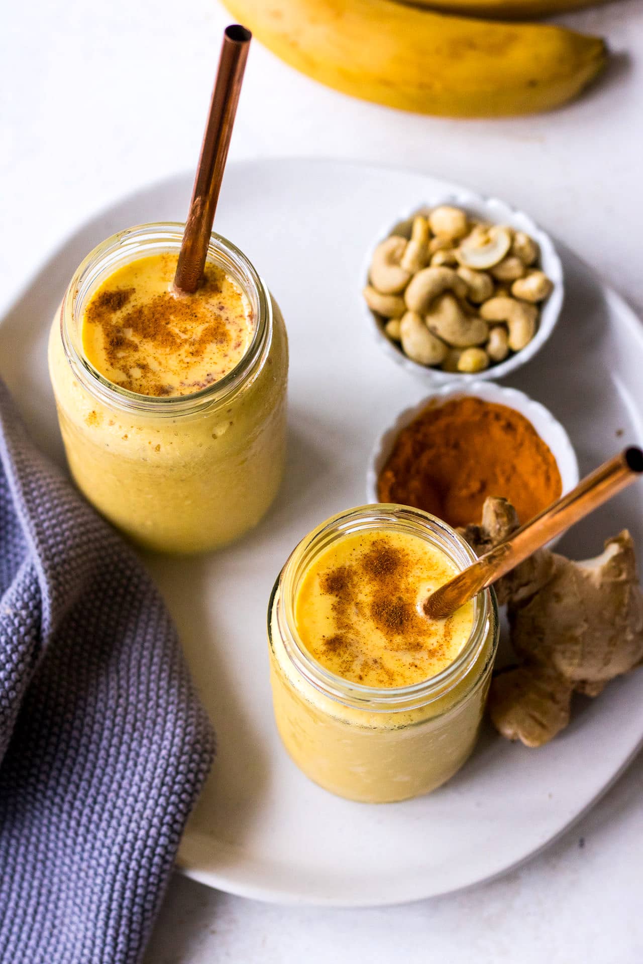 Two glass jars filled with a turmeric golden milk smoothie with copper straws, cashews and turmeric in background
