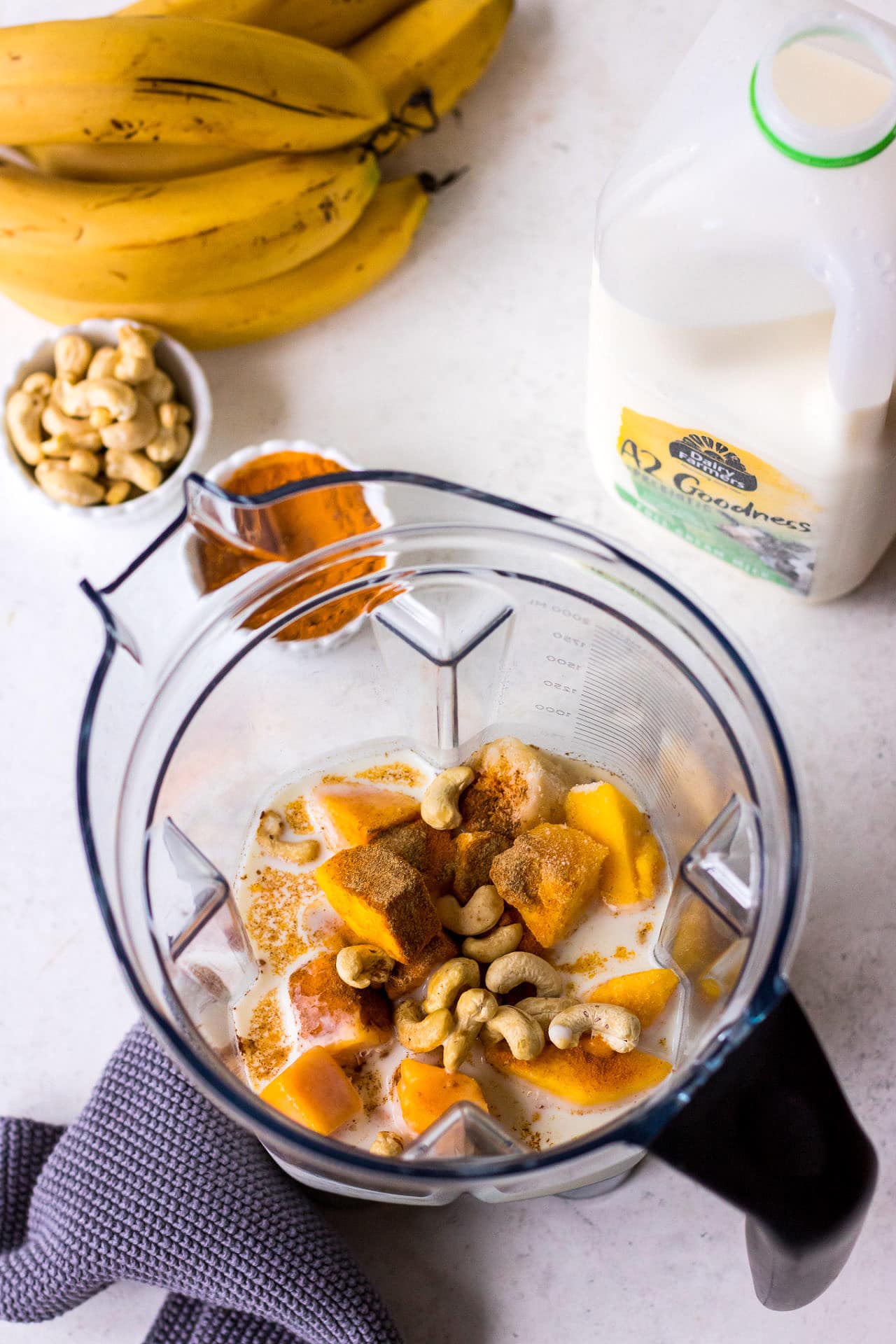 Blender jug filled with mango, turmeric, cashews and milk with milk carton and cashews in background