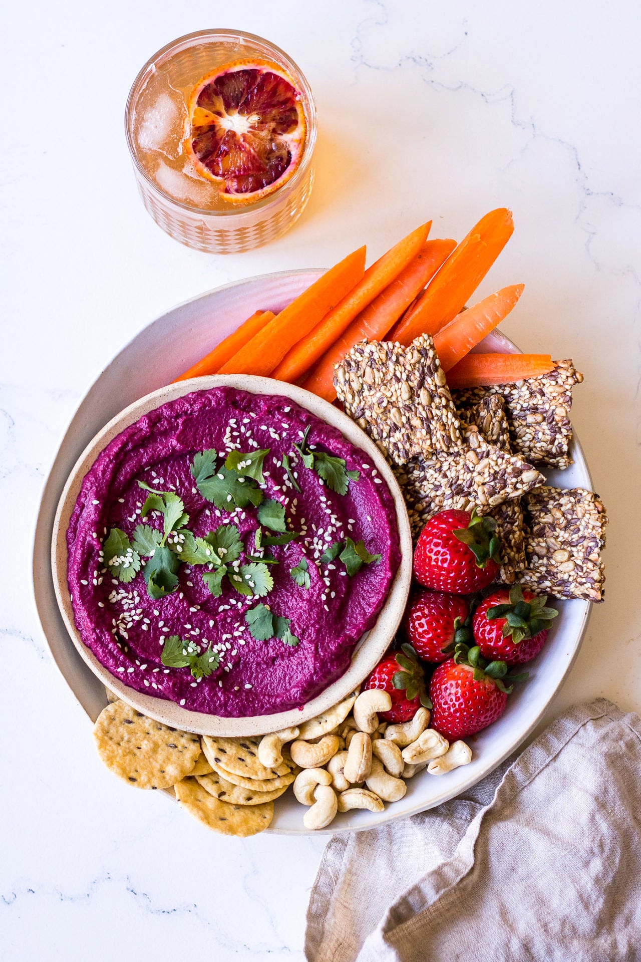 round dish of beet dip arranged on a ceramic plate with crackers, strawberries and carrot sticks
