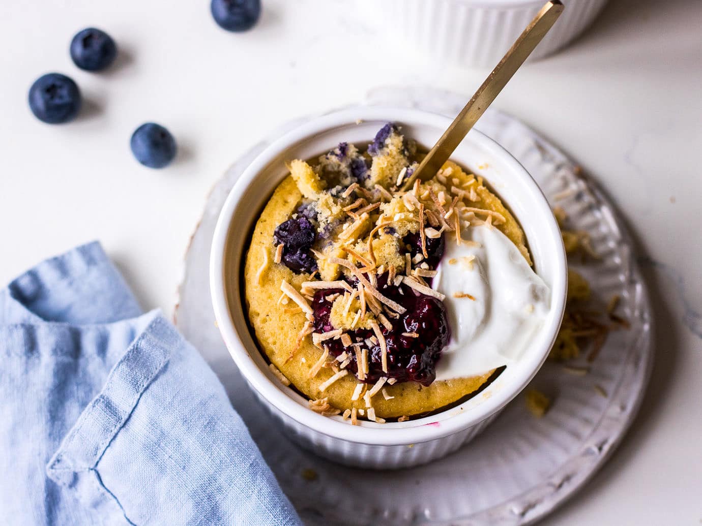Vanilla cake in white ramekin topped with blueberries, yoghurt and toasted coconut, gold spoon sticking out