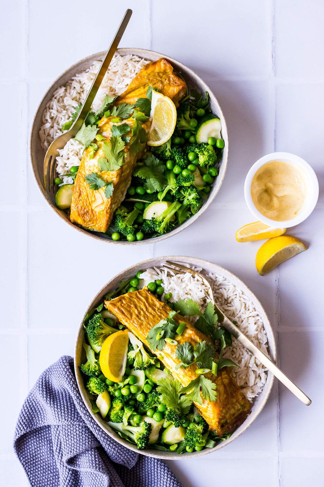 Two bowls with white rice, green vegetables and curry baked salmon garnished with lemon and coriander