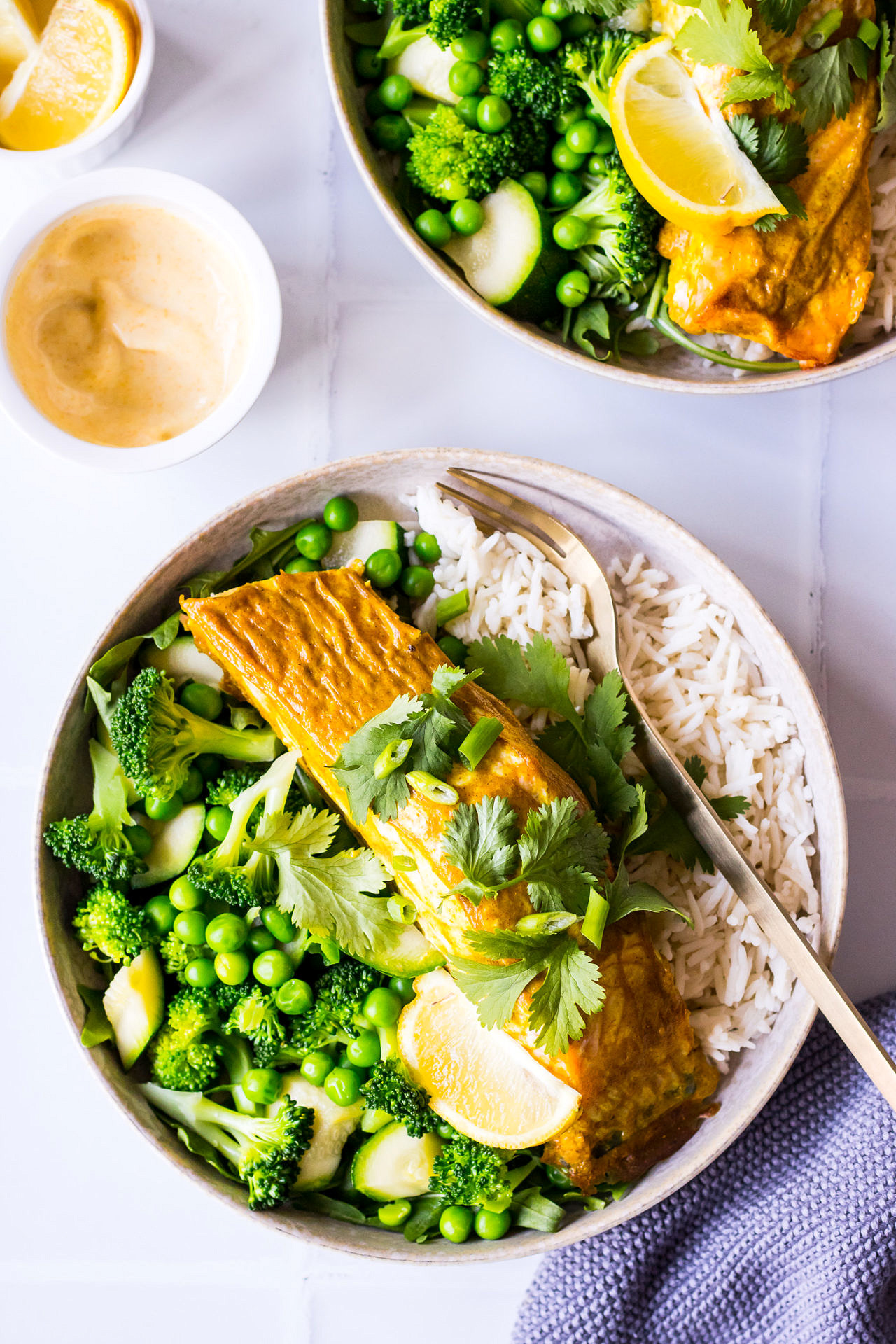 Salmon fillet on top of white rice and green vegetables garnished with coriander, spiced yoghurt in small dish to the side