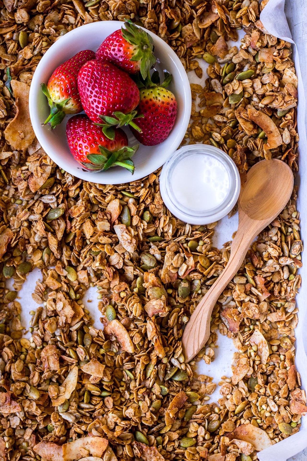 Baking tray spread out with granola, small bowl of strawberries, milk jar and small wooden spoon