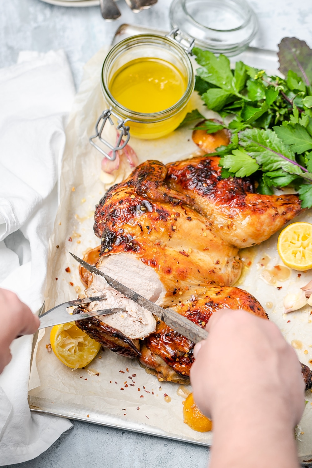 Hands carving into one side of a flattened roast chicken surrounded by lemons, pan juices and fresh herbs