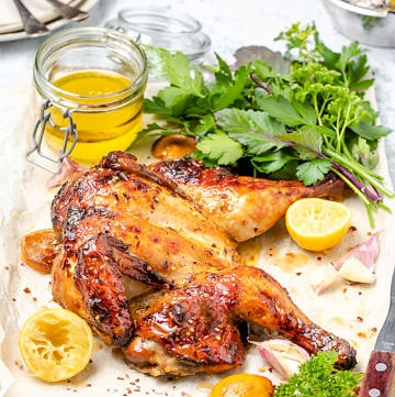 Butterflied roast chicken surrounded by lemons, fresh herbs and small jar of pan juices