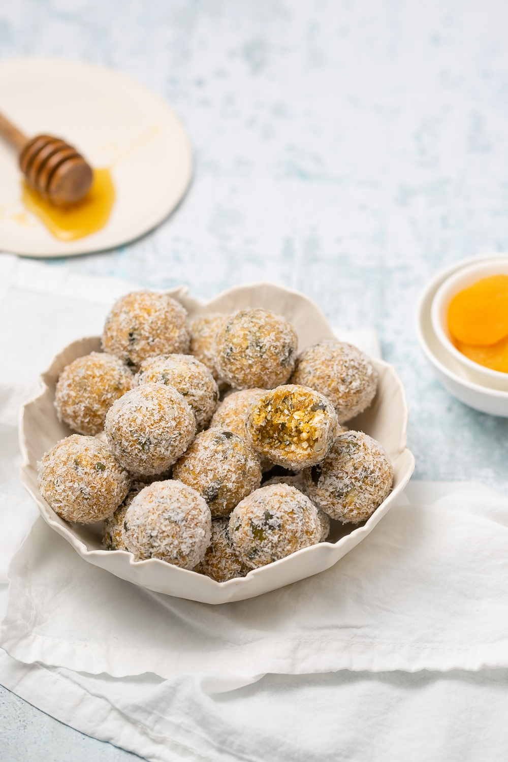 small white scalloped bowl filled with apricot bliss balls, white napkin and honey to side