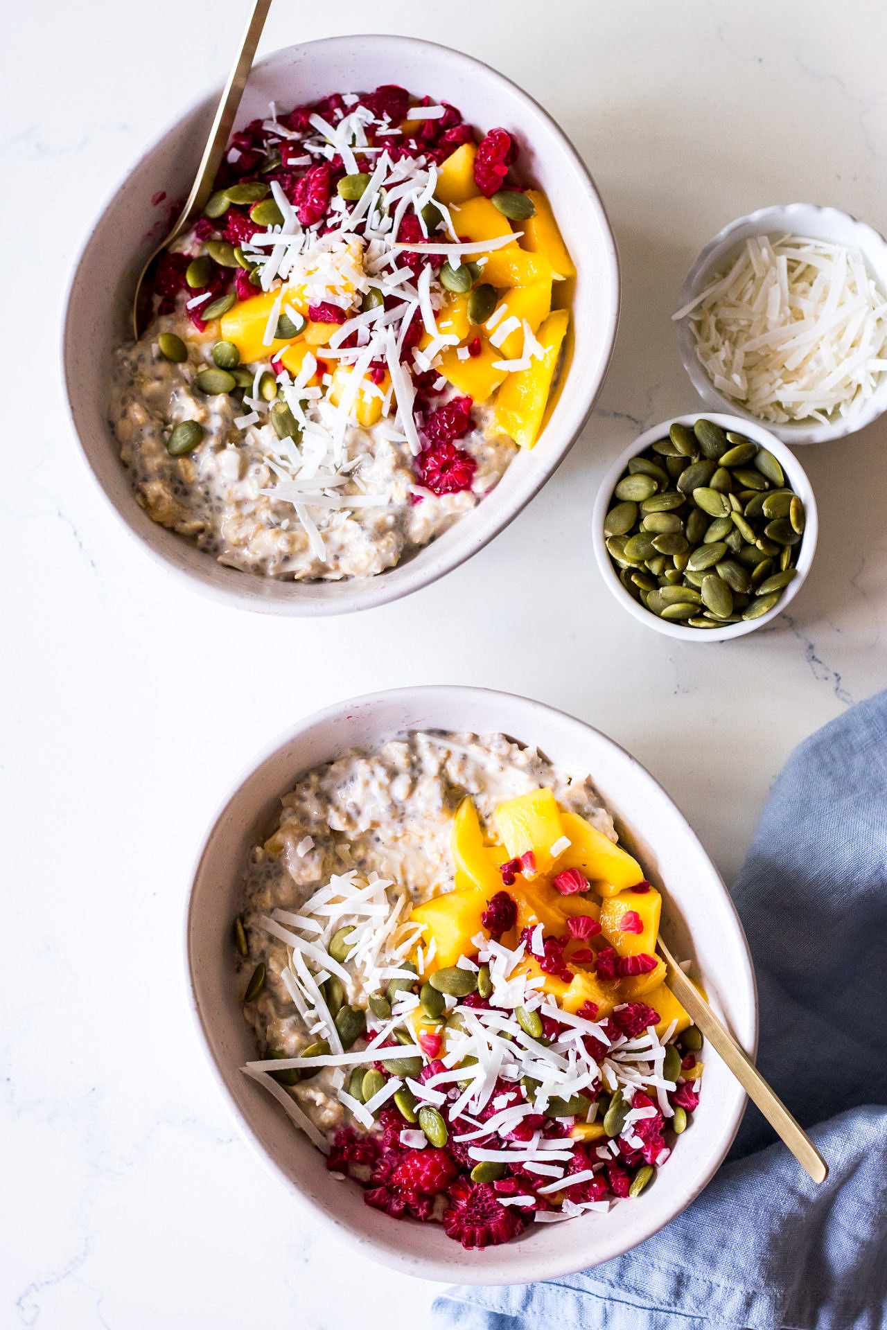 Top view of two white ceramic bowls of overnight chia oats with mango, coconut and raspberries