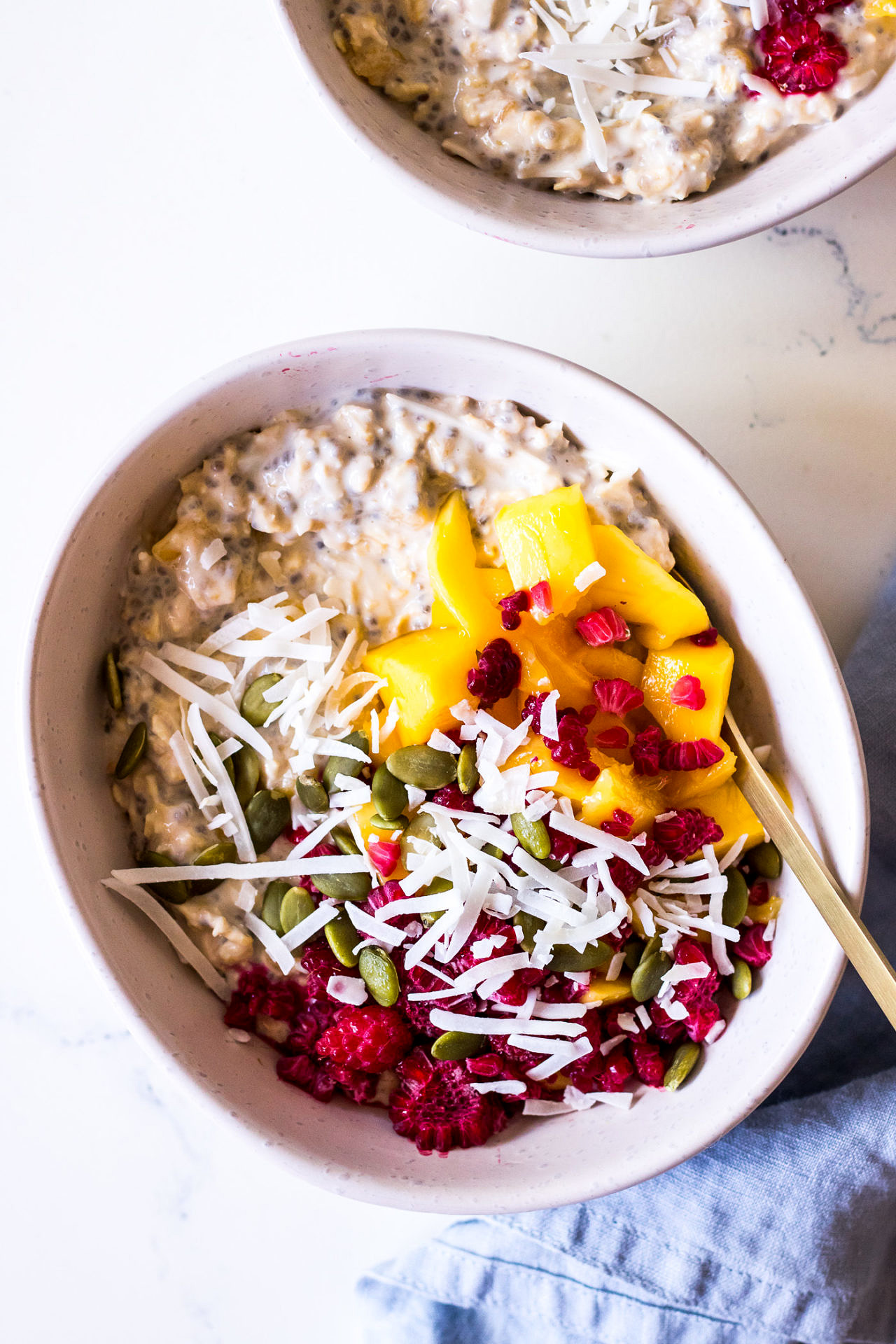 White bowl of overnight oats with chia seeds, mango, shredded coconut and raspberries
