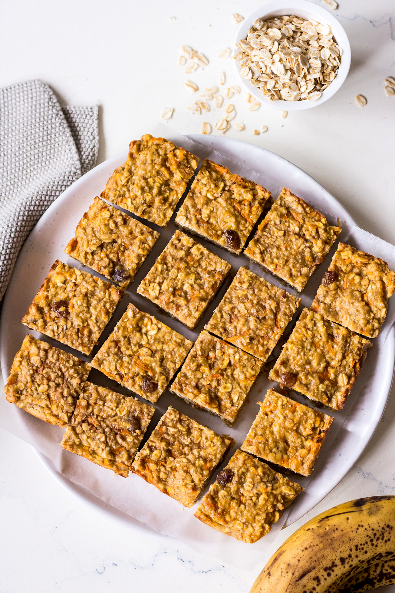 Oatmeal bars arranged on a white ceramic plate with rolled oats and banana