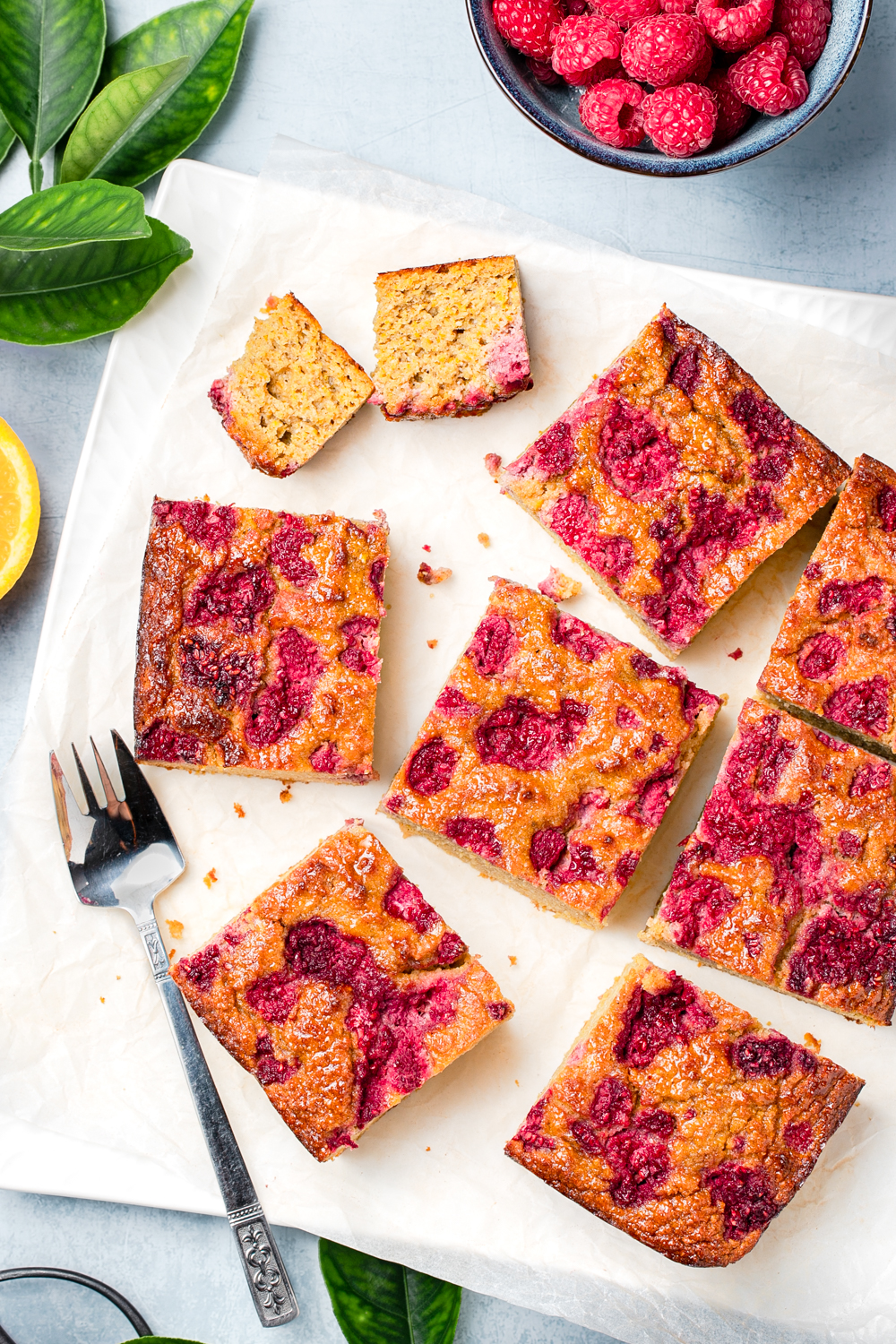 Square slices of orange almond and raspberry healthy cake against a blue background with fresh raspberries