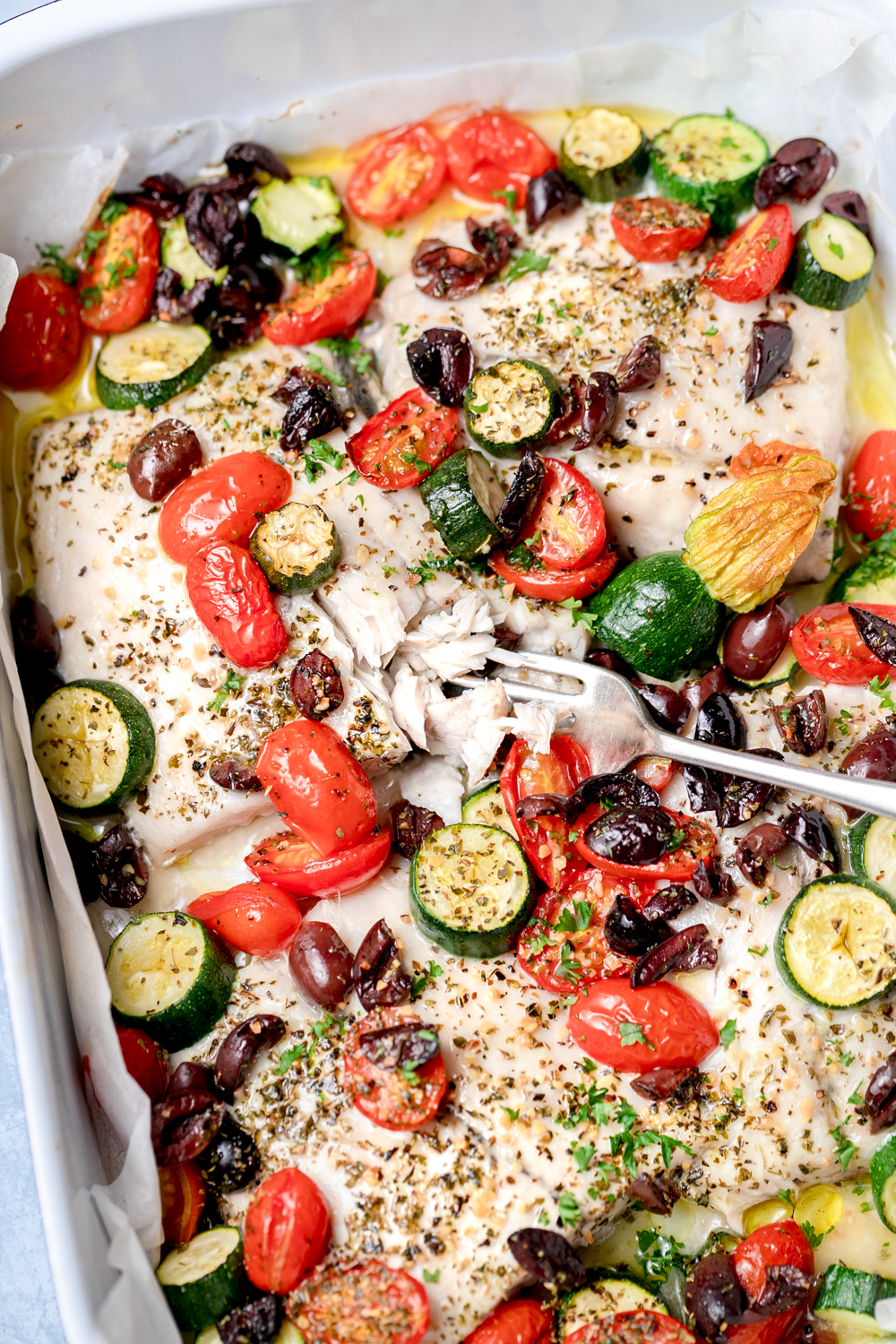 Close up baked white fish fillets with cherry tomatoes, zucchini, olives and herbs