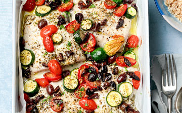 Fish tray bake with zucchini, tomato and olives on a blue background