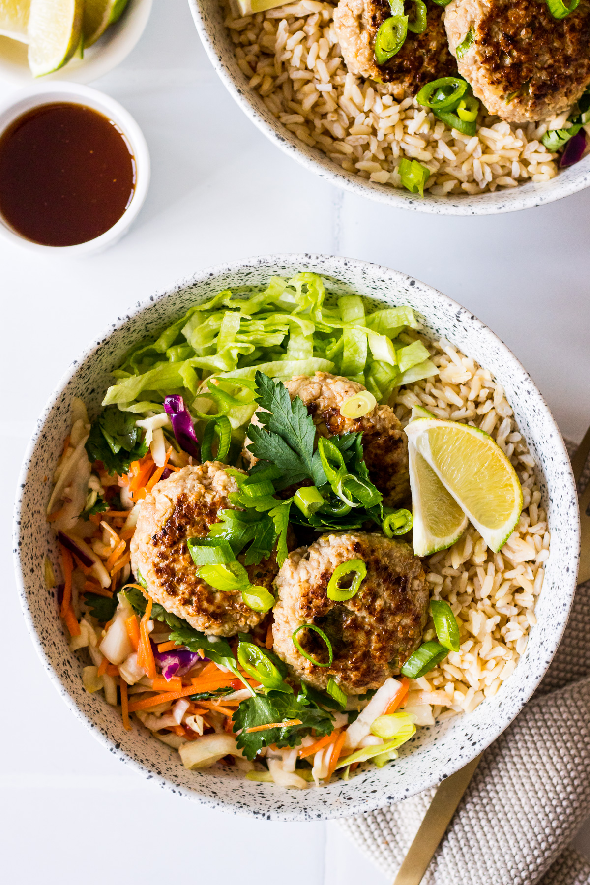 Speckled white bowl with brown rice, lemongrass pork patties, salad, spring onion, lime wedge