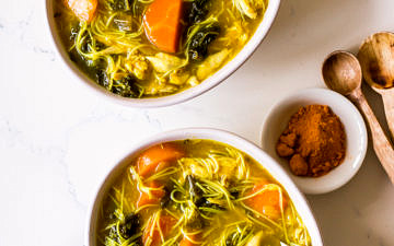 Two bowls of turmeric chicken noodle soup with gold spoons and a blue napkin
