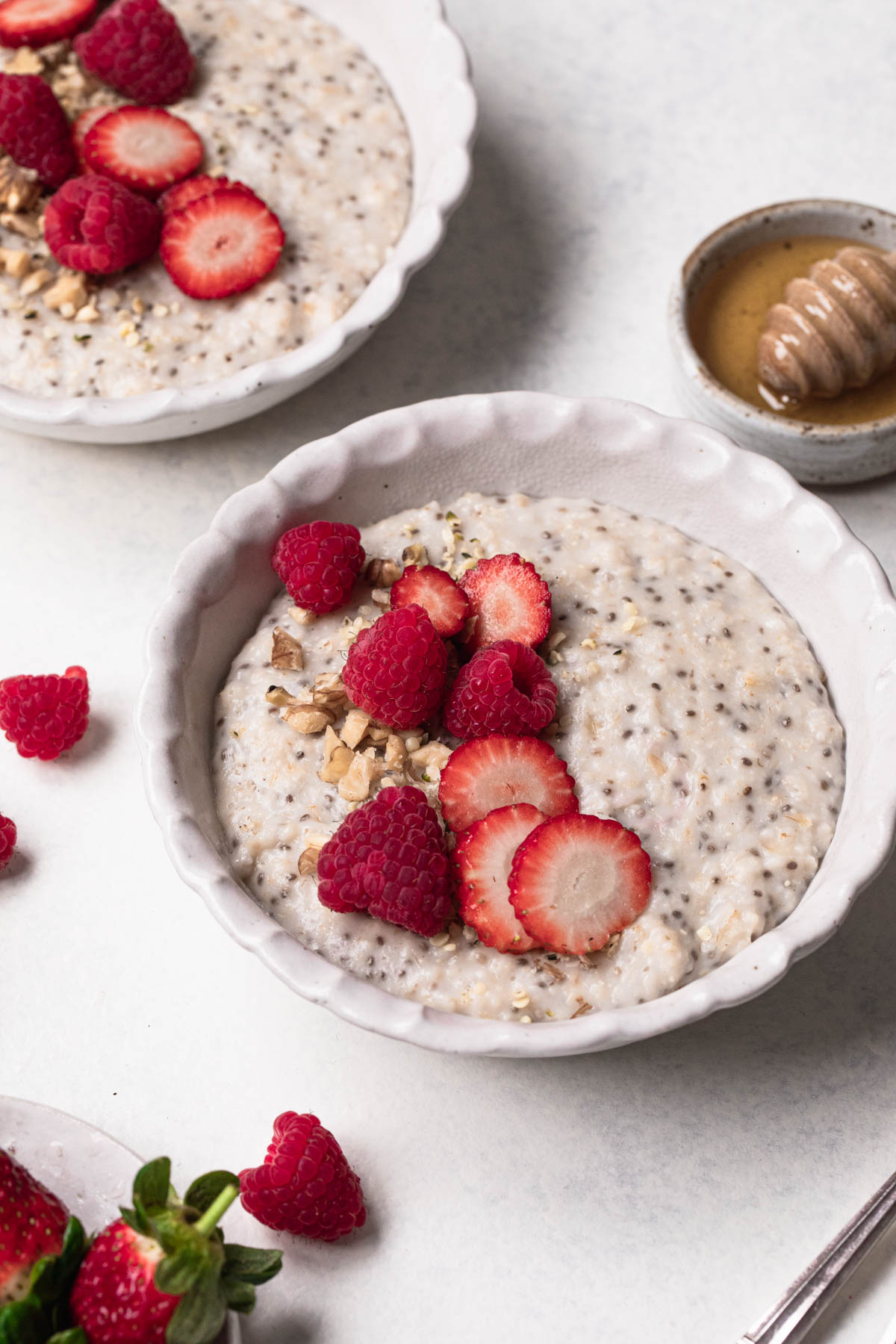 Two bowls of porridge with chia seeds, sliced strawberries and honey on a white table