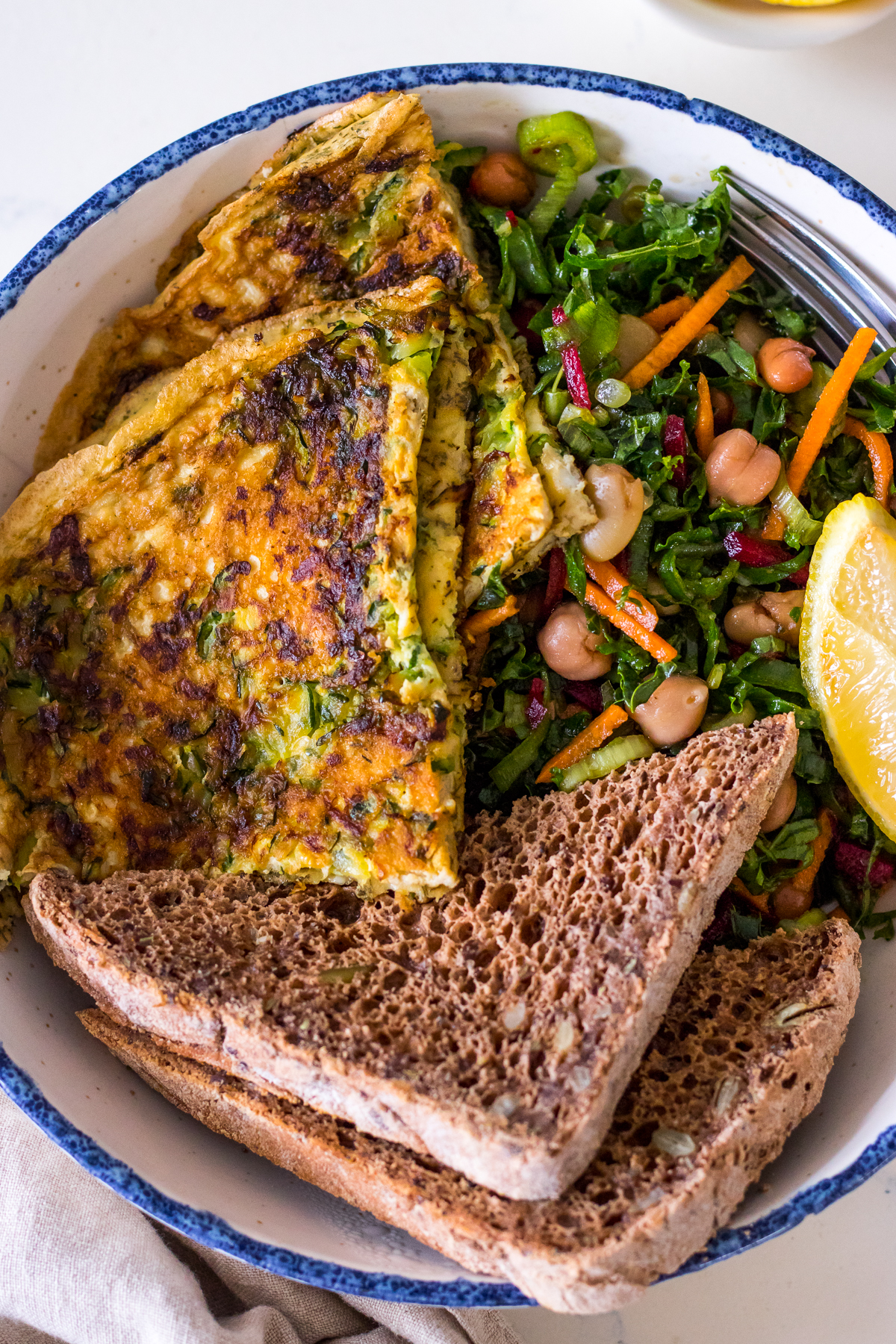 Zucchini omelette with dried herbs in a white bowl with kale slaw and brown seeded toast