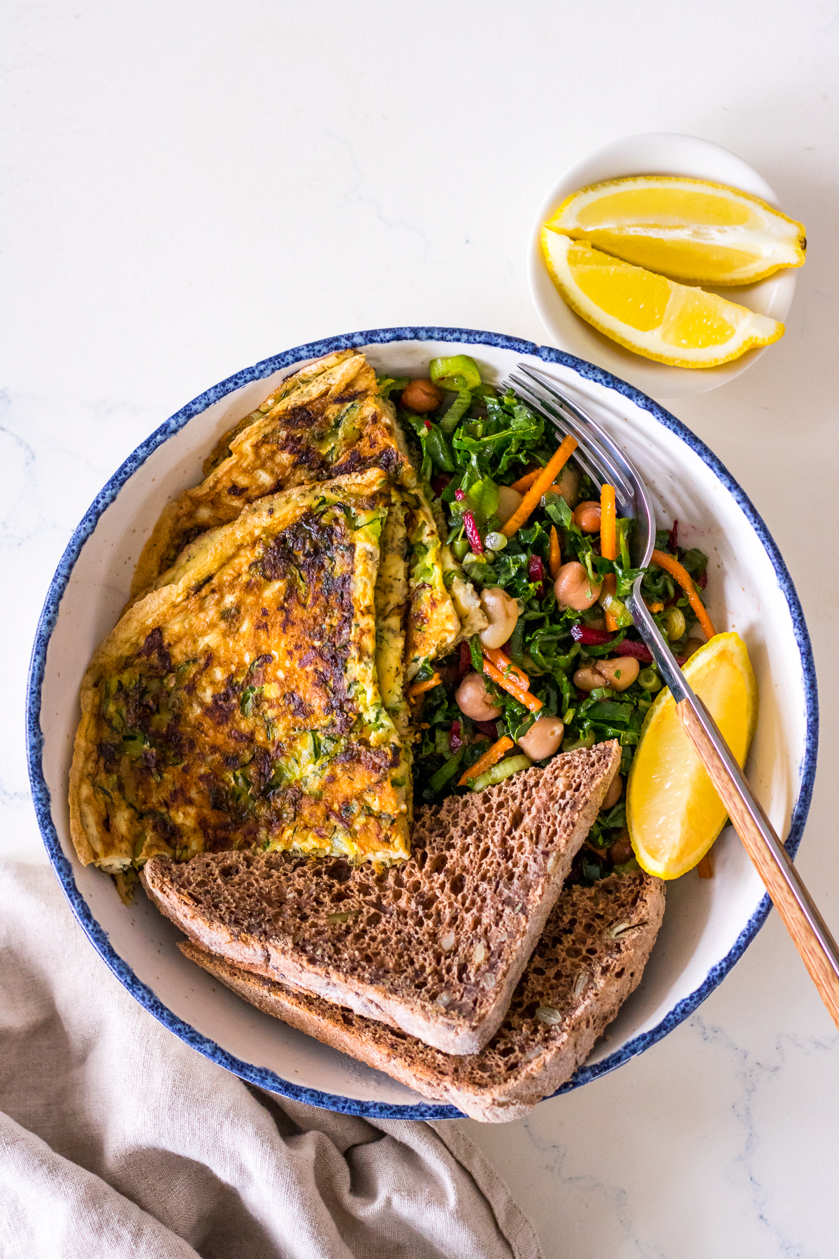 Shallow bowl with zucchini omelette, seeded toast and kale salad, lemon wedge