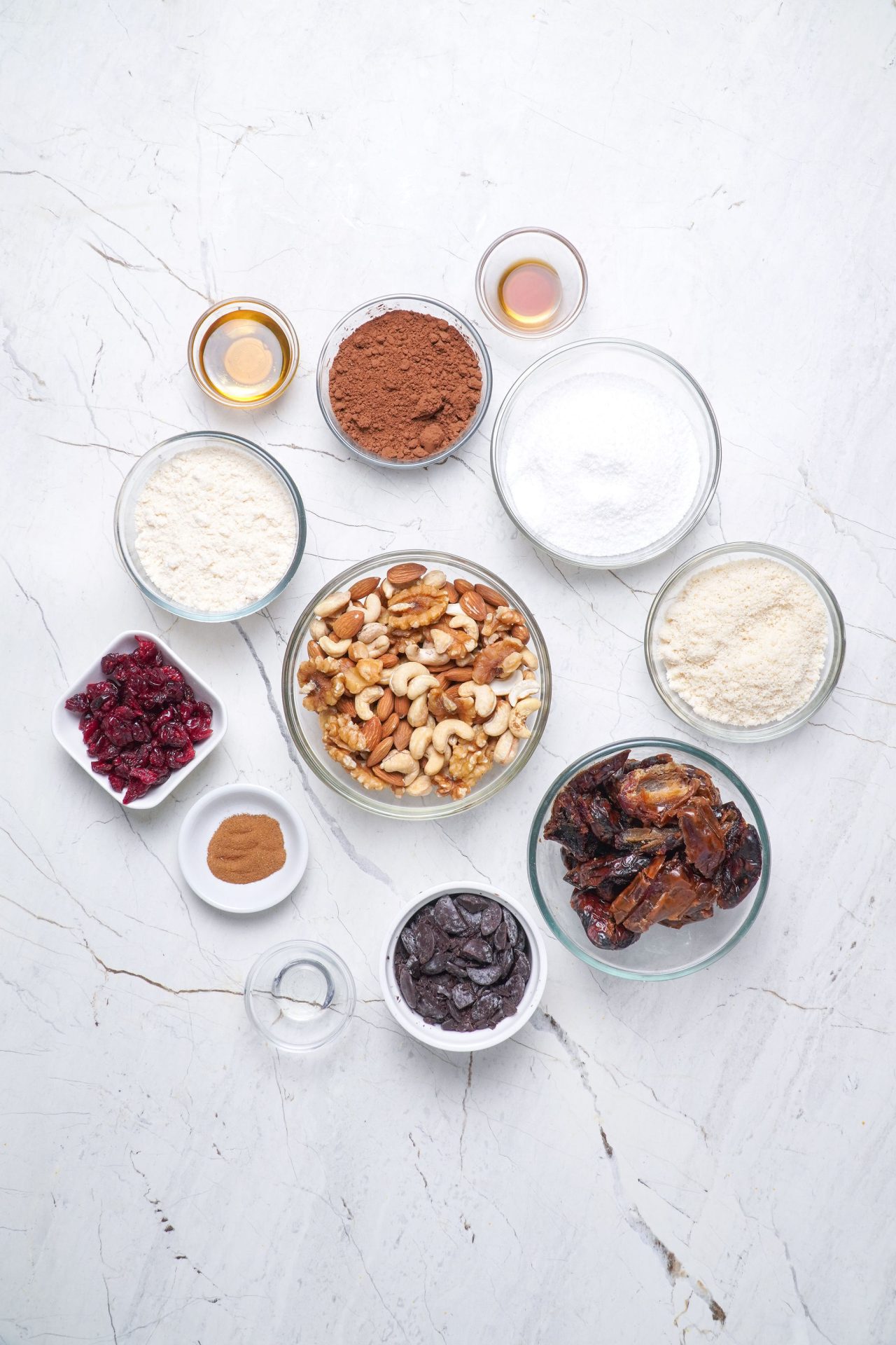 Ingredients to make healthy truffles. In small bowls. Honey, cacao powder, medjool dates, coconut flour, mixed nuts, cranberries, cinnamon, vanilla, dark chocolate.
