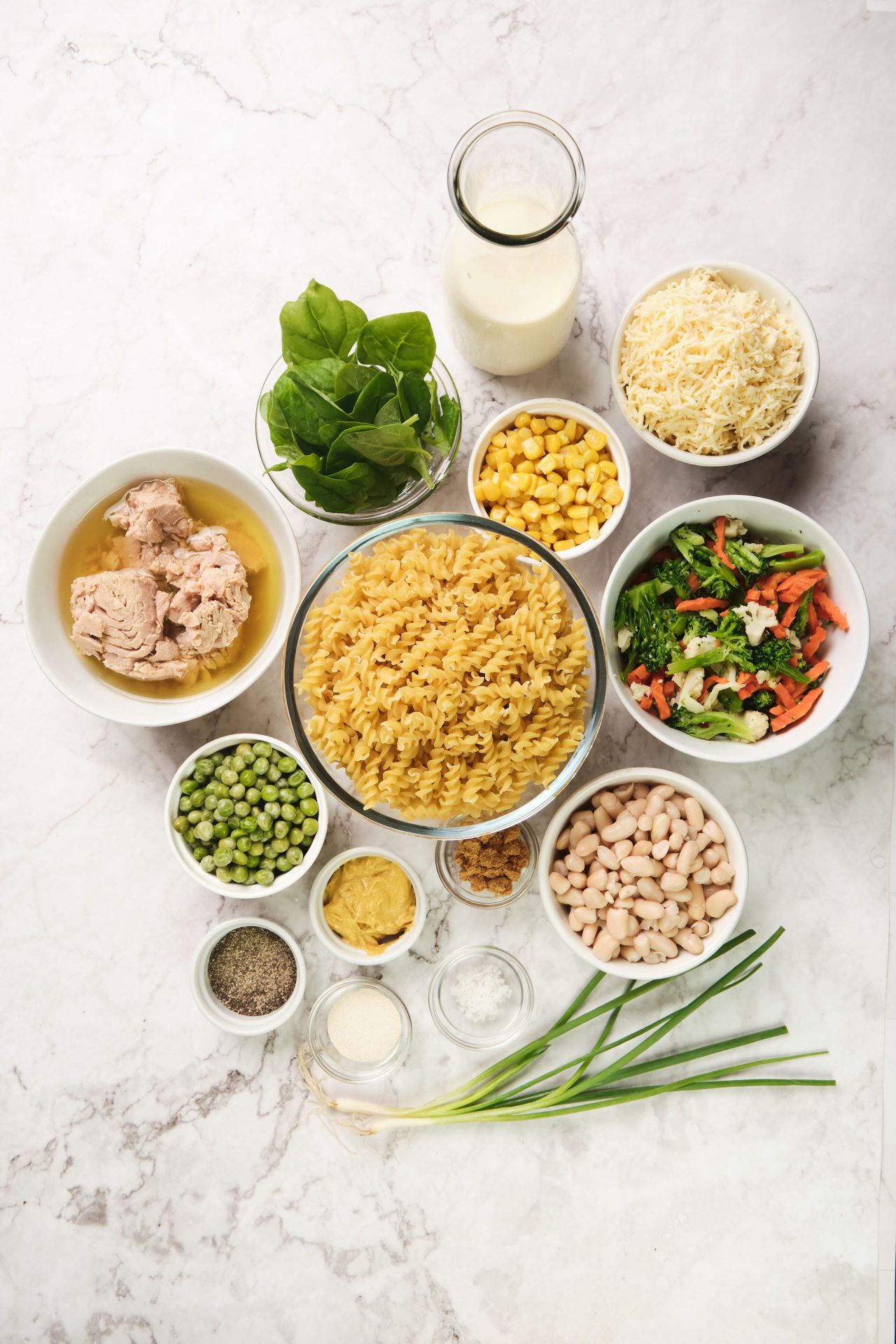 Flatlay of ingredients to make tuna pasta bake including dried spiral pasta, canned tuna, frozen vegetables, milk, cannellini beans and shredded cheese