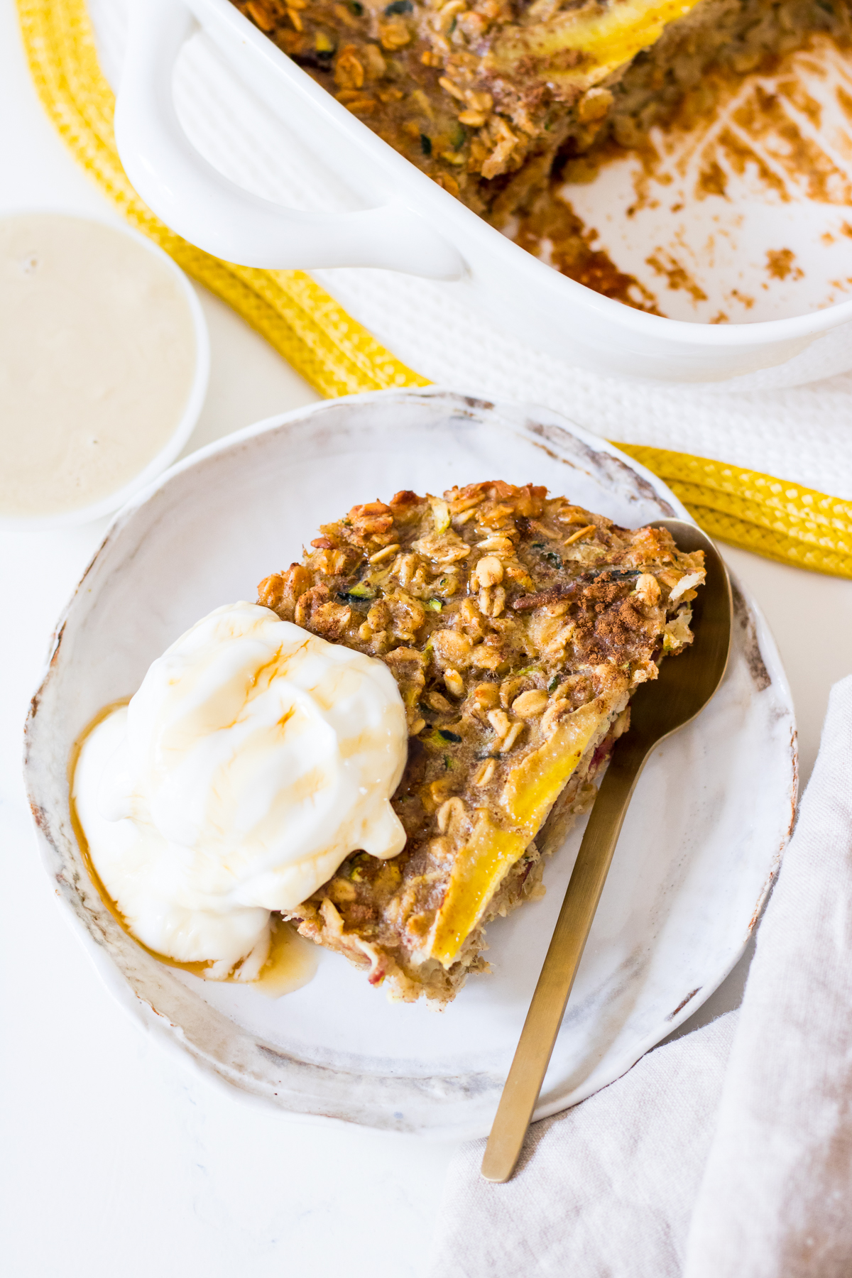 A slice of banana zucchini baked oatmeal on a white side plate with yoghurt