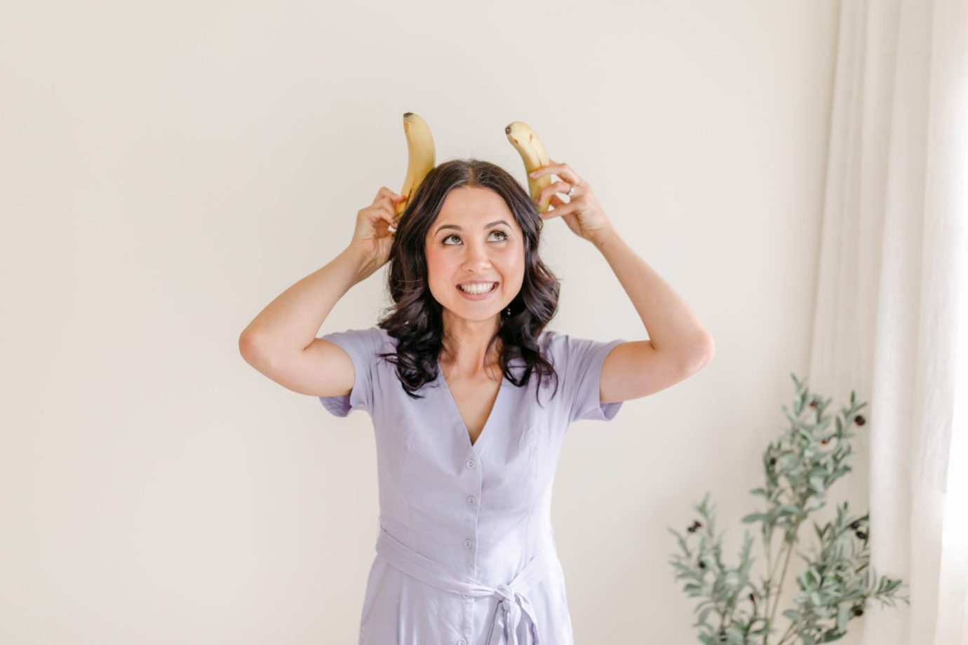Monique Cormack blogger at Nourish Everyday, wearing a purple dress and holding bananas up to her head, laughing